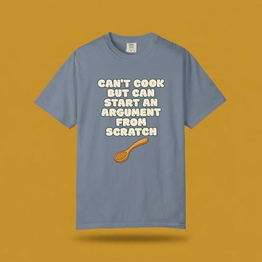 Blue graphic t-shirt flat lay on yellow background featuring Can’t Cook slogan.