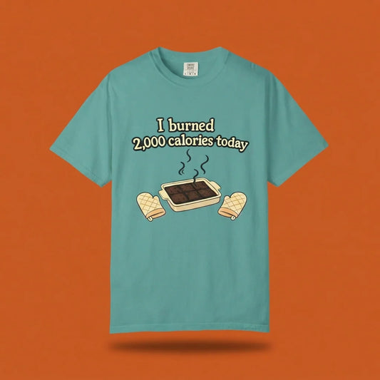 A teal garment-dyed Comfort Colors t-shirt displayed flat, featuring a humorous graphic of a smoking brownie pan and the sarcastic phrase "I burned 2,000 calories today."