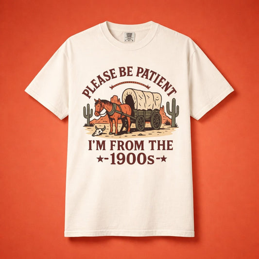 Flat lay of a cream Comfort Colors t-shirt featuring a vintage covered wagon illustration and text Please Be Patient I'm From The 1900s.