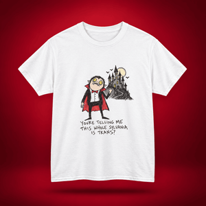 A white short-sleeve crewneck t-shirt featuring a cartoon graphic of a vampire looking surprised next to a haunted castle, with the text 'YOU'RE TELLING ME THIS WHOLE SILVANIA IS TRANS?' printed below. The t-shirt is displayed against a dark red gradient background.