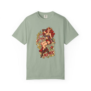 A sage green short-sleeve t-shirt with a vintage-style pin-up graphic on the front. The graphic features a woman with red hair and a red dress, surrounded by retro text that reads 'ANYTHING CAN BE A MIMIC'.