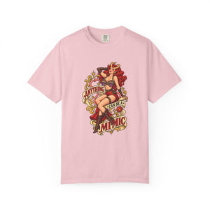 A light pink short-sleeve crewneck t-shirt featuring a vintage-style pin-up graphic on the front. The graphic depicts a woman with red hair in a red and black outfit, surrounded by decorative elements and the text 'ANYTHING CAN BE A MIMIC.