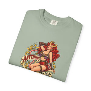 A folded sage green t-shirt featuring a colorful retro pin-up style graphic of a horned woman with the text 'ANYTHING CAN BE A MIMIC' on a white background.