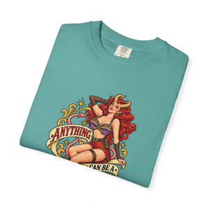A folded teal t-shirt with a colorful graphic print of a pin-up style warrior woman with red hair and horns, sitting on a banner that reads 'ANYTHING I CAN BE A MIMIC'.