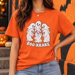 A woman wearing an orange graphic t-shirt with a cartoon ghost design and the text 'BOO-KKAKE', paired with blue jeans, set against a Halloween-themed background with pumpkins and a decorated wall.