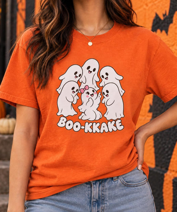 A woman wearing an orange graphic t-shirt with a cartoon ghost design and the text 'BOO-KKAKE', paired with blue jeans, set against a Halloween-themed background with pumpkins and a decorated wall.