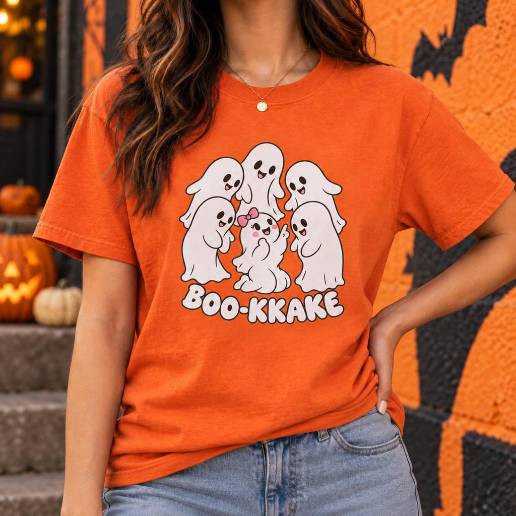 A woman wearing an orange graphic t-shirt with a cartoon ghost design and the text 'BOO-KKAKE', paired with blue jeans, set against a Halloween-themed background with pumpkins and a decorated wall.