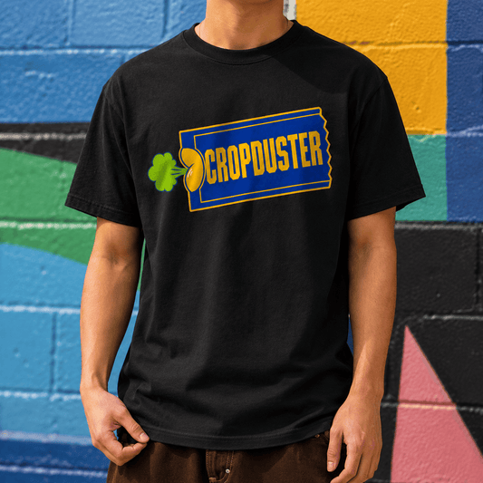 Flat lay mockup of the black Cropduster pop culture joke shirt centered on a vibrant royal blue gradient background.