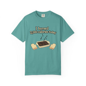 Flat lay view of a teal-colored t-shirt with a sarcastic baking graphic showing overdone brownies and the phrase "I burned 2,000 calories today."