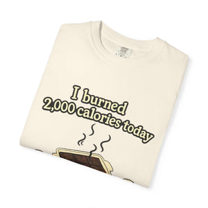A folded ivory or cream-colored t-shirt with a humorous illustration of smoking brownies and the text "I burned 2,000 calories today."