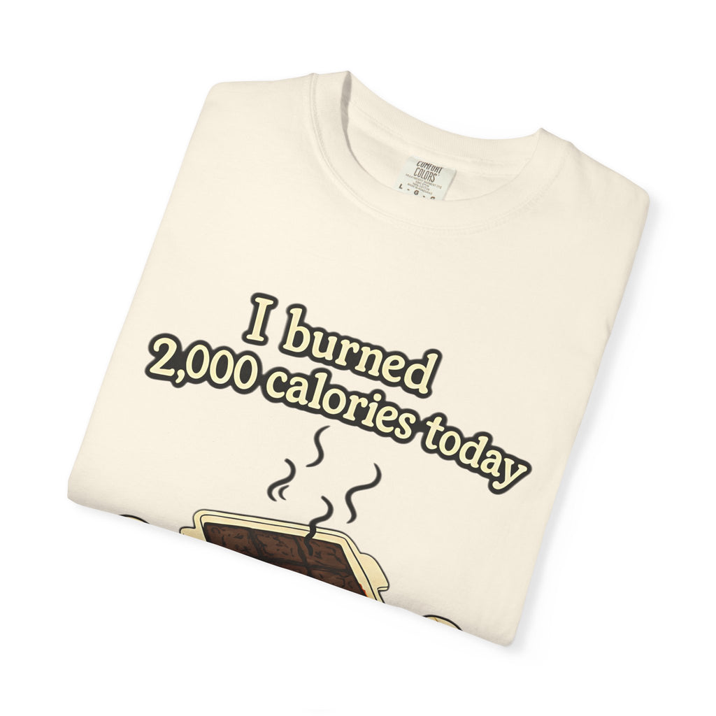 A folded ivory or cream-colored t-shirt with a humorous illustration of smoking brownies and the text "I burned 2,000 calories today."