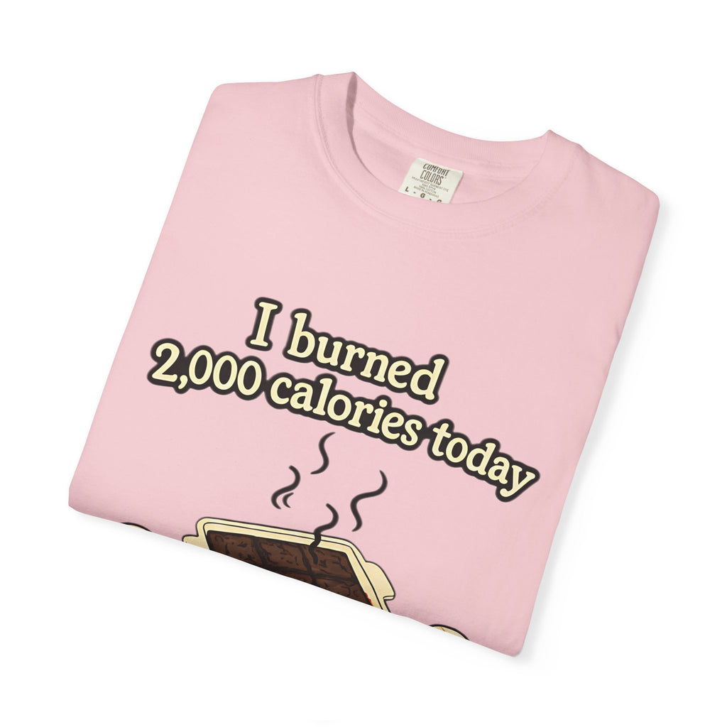 A light pink folded t-shirt featuring a funny cartoon graphic of a baking fail and the caption "I burned 2,000 calories today."