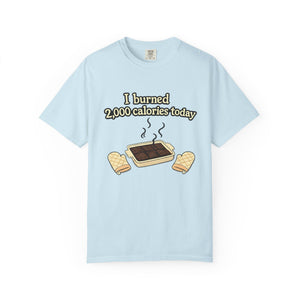 A flat lay of a light blue t-shirt featuring a funny "I burned 2,000 calories today" baking meme with a smoking brownie pan graphic.