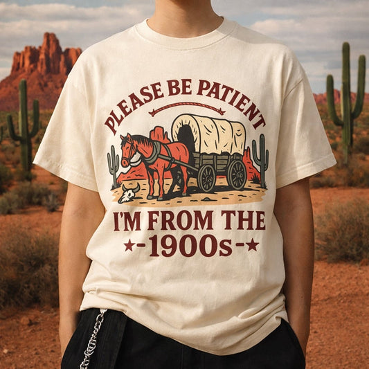 Flat lay of a cream Comfort Colors t-shirt featuring a vintage covered wagon illustration and text Please Be Patient I'm From The 1900s.