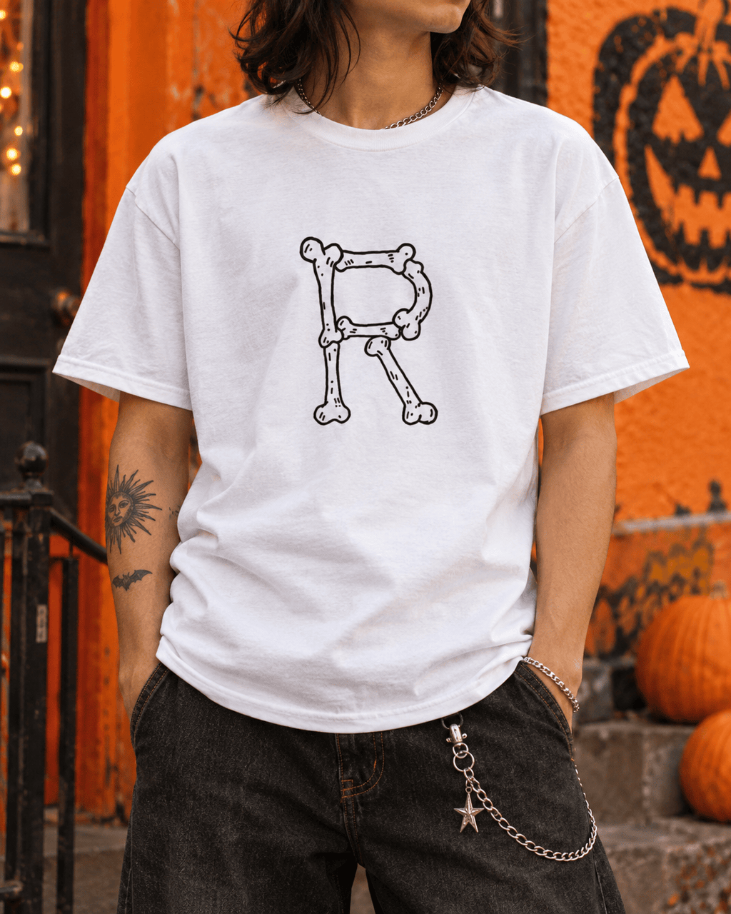 A person wearing a white graphic T-shirt with a black skeletal letter 'R' on the chest, paired with dark jeans, standing in front of an orange Halloween-themed wall with pumpkins.