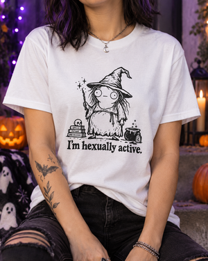 A person wearing a white graphic T-shirt with a black witch illustration and the text 'I'm hexually active.' The photo is styled in a Halloween-themed setting with pumpkins, candles, and dark ambient background lighting.