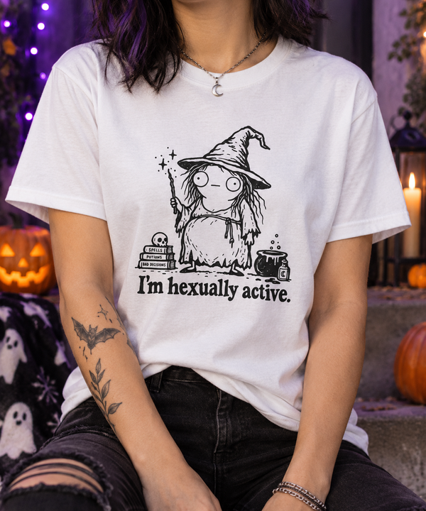 A person wearing a white graphic T-shirt with a black witch illustration and the text 'I'm hexually active.' The photo is styled in a Halloween-themed setting with pumpkins, candles, and dark ambient background lighting.