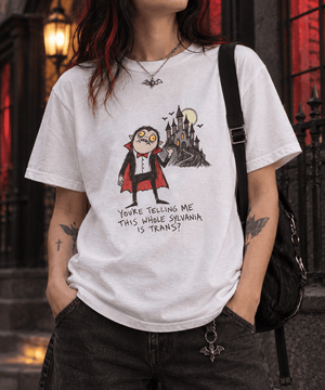 A person wearing a white graphic T-shirt with a gothic vampire and castle print, paired with dark jeans and a black shoulder bag, set against a moody gothic doorway background.
