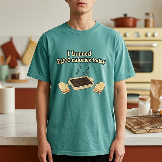 A teal garment-dyed Comfort Colors t-shirt displayed flat, featuring a humorous graphic of a smoking brownie pan and the sarcastic phrase "I burned 2,000 calories today."