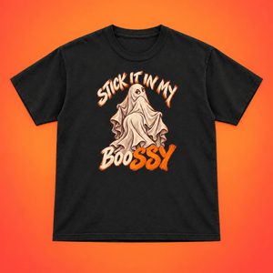 A black short-sleeve t-shirt with a Halloween graphic featuring a ghost and the text 'STICK IT IN MY BOOSSY' in orange and beige, displayed flat against a vibrant orange-to-yellow gradient background.