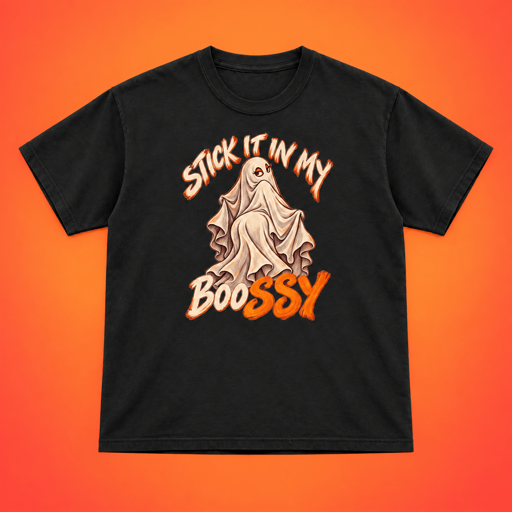A black short-sleeve t-shirt with a Halloween graphic featuring a ghost and the text 'STICK IT IN MY BOOSSY' in orange and beige, displayed flat against a vibrant orange-to-yellow gradient background.