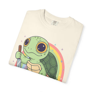 Flat lay of a folded ivory Comfort Colors t-shirt featuring a cartoon turtle holding a knife with a rainbow background.