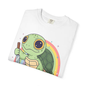 Flat lay of a folded white Comfort Colors t-shirt featuring a cartoon turtle holding a knife with a rainbow background.